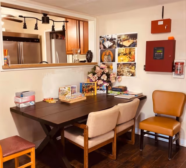 A small room with a dark wooden table surrounded by four chairs, two beige cushioned chairs, one brown leather chair, and one wooden stool. On the table are various board games, books, and a vase with pink flowers. The wall behind the table has a fire alarm panel, a fire extinguisher, and several photos pinned up. A pass-through window reveals a kitchen area with stainless steel appliances and wooden cabinets.