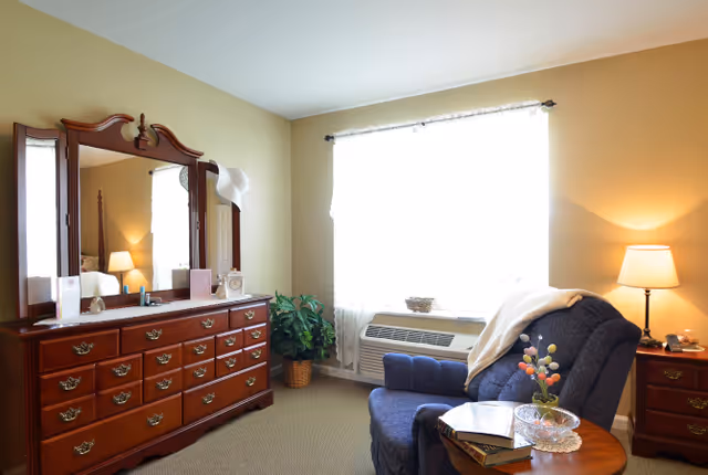 Sunlit bedroom with a wooden dresser and mirror, a blue upholstered armchair beside a small table and lamp, and a window air conditioner.
