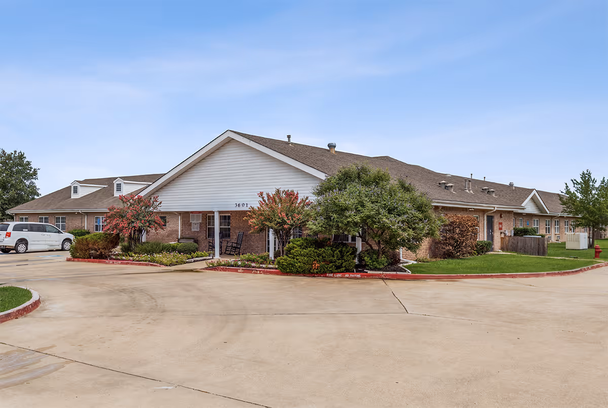 Exterior view of Bluebonnet Court, a single-story brick building with a white gabled roof and landscaped greenery including bushes and small trees. A concrete driveway and parking area are visible in front of the building under a clear blue sky.