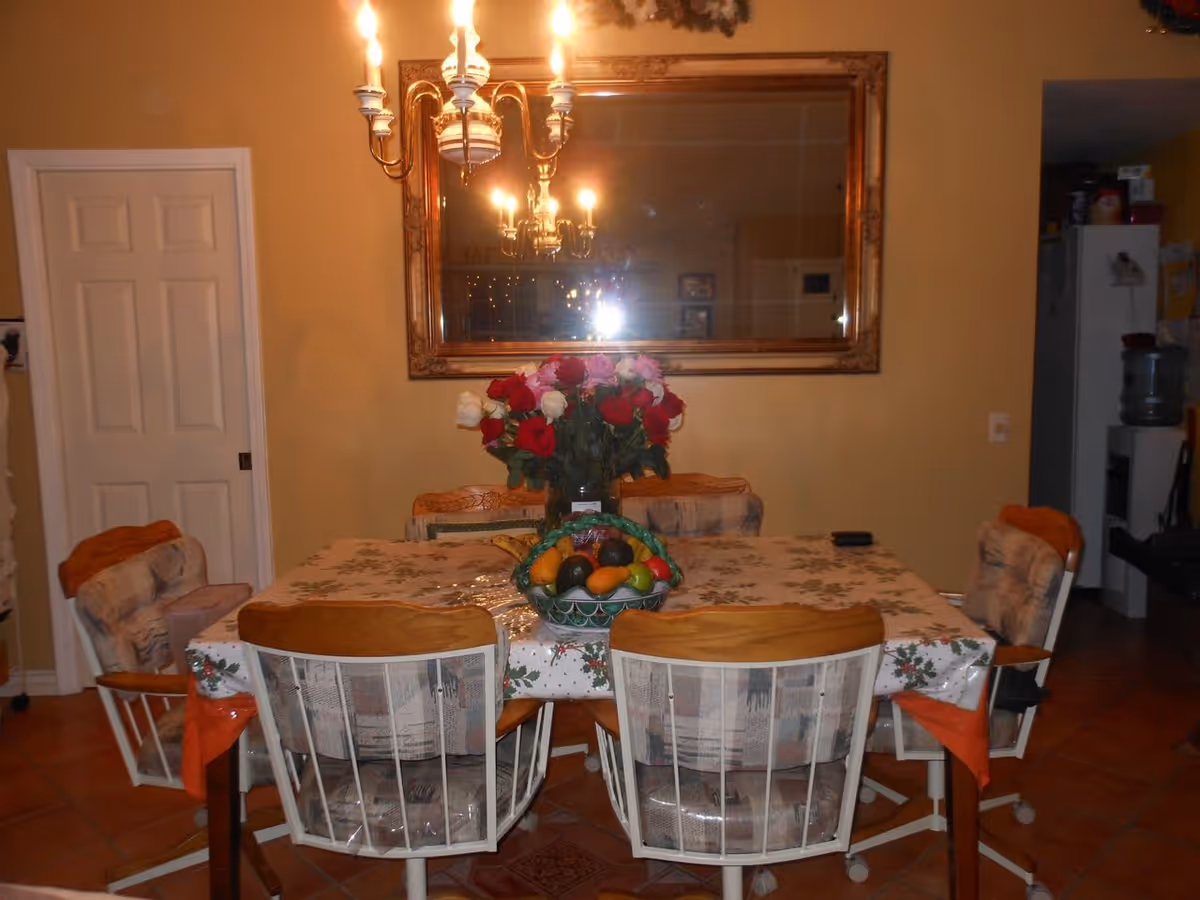 A dining room with a rectangular table covered with a floral tablecloth. Six cushioned chairs with wooden backs surround the table. On the table, there is a basket of assorted fruits and a vase with red, pink, and white roses. A large ornate mirror hangs on the wall behind the table, reflecting a chandelier with lit bulbs. The room has warm yellow walls and a white door on the left side.