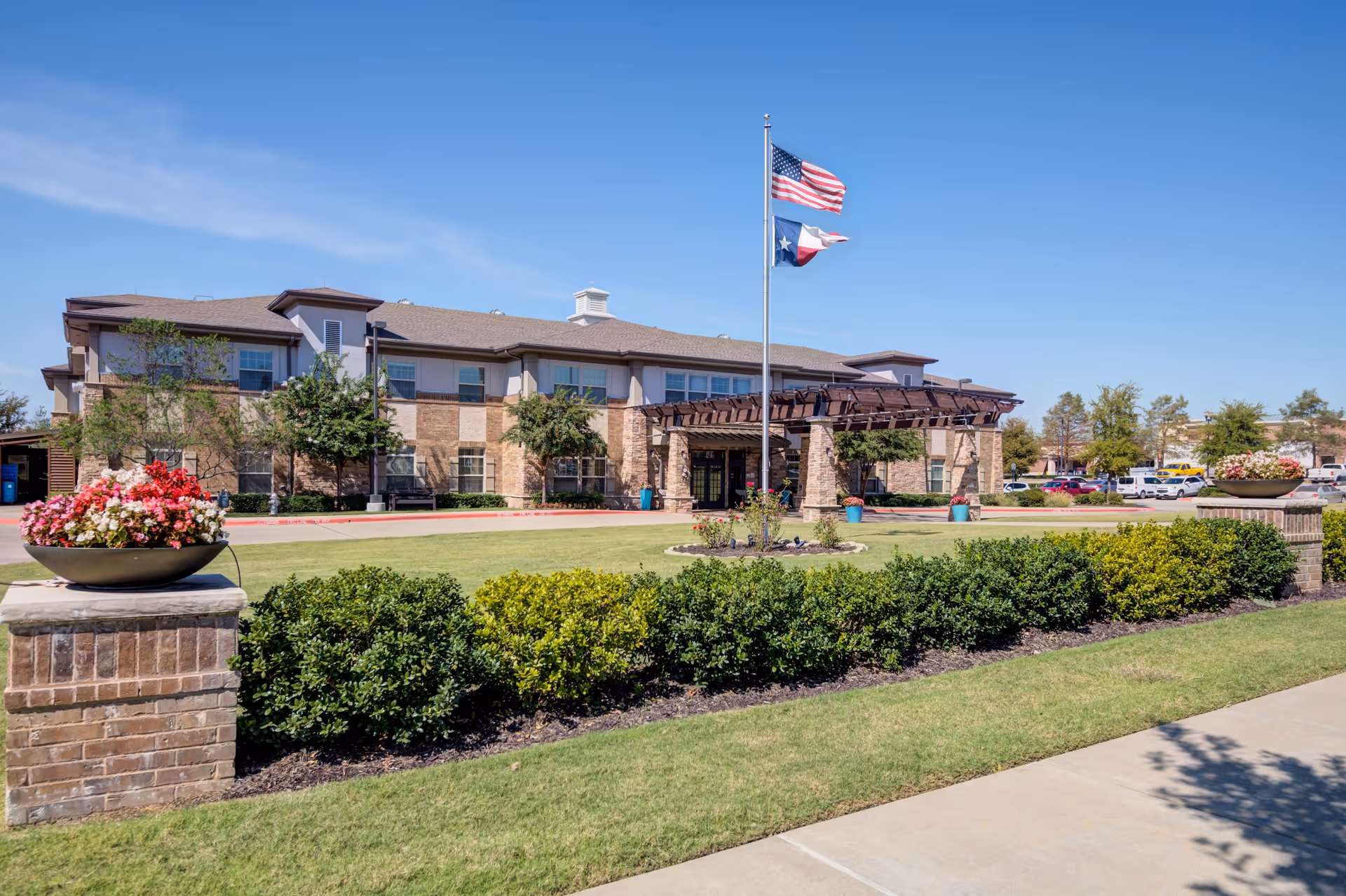 Exterior view of Truewood by Merrill, Keller senior living facility showing a two-story building with a covered entrance, American and Texas flags on a flagpole, landscaped bushes, flower pots, and a clear blue sky.