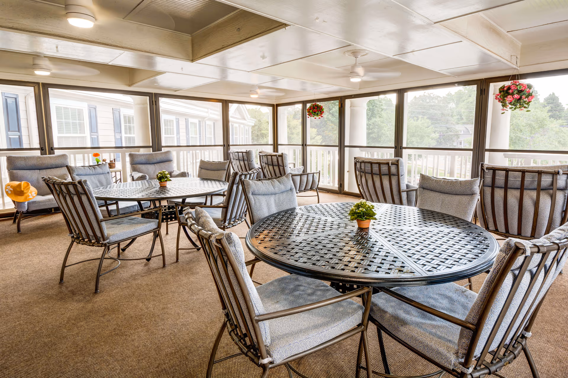 A bright enclosed patio area with multiple round metal tables and cushioned chairs arranged around them. The space has large windows with white frames, hanging flower pots, ceiling fans, and a carpeted floor. Outside, greenery and parts of a building are visible through the windows.
