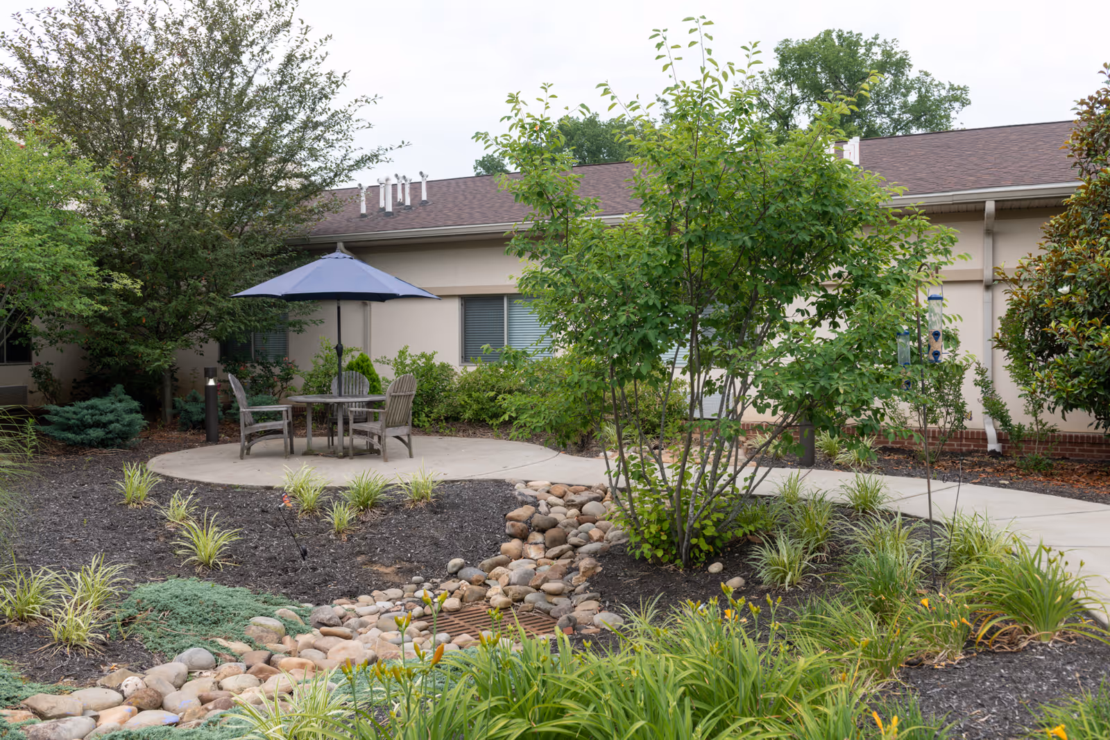 Outdoor courtyard featuring a circular patio with a table, chairs and a blue umbrella, bordered by landscaping and a low single-story building.