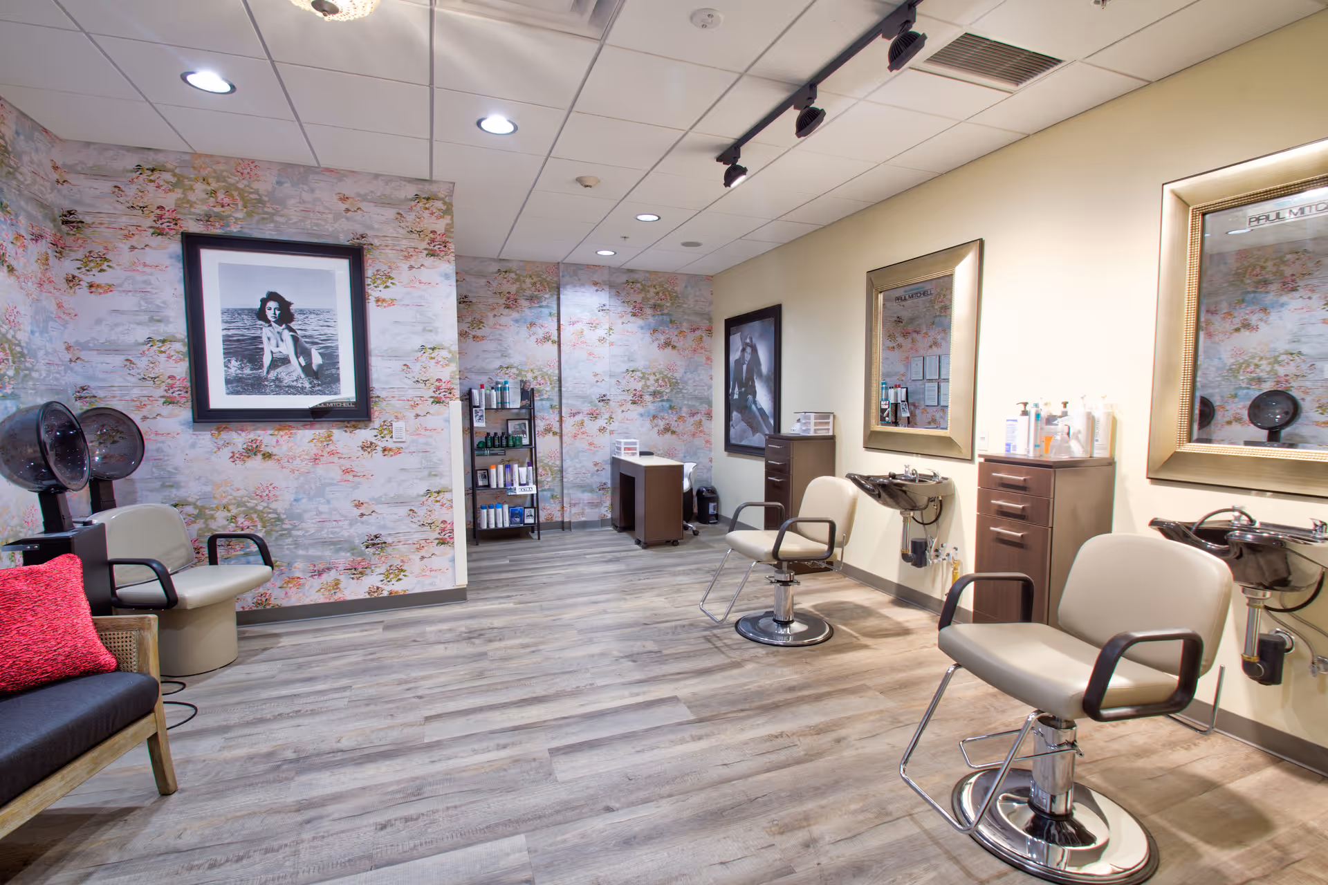 Interior view of a salon area with two salon chairs in front of mirrors and sinks, a waiting chair with a red pillow, hair dryers, floral wallpaper on one wall, and shelves with hair products.