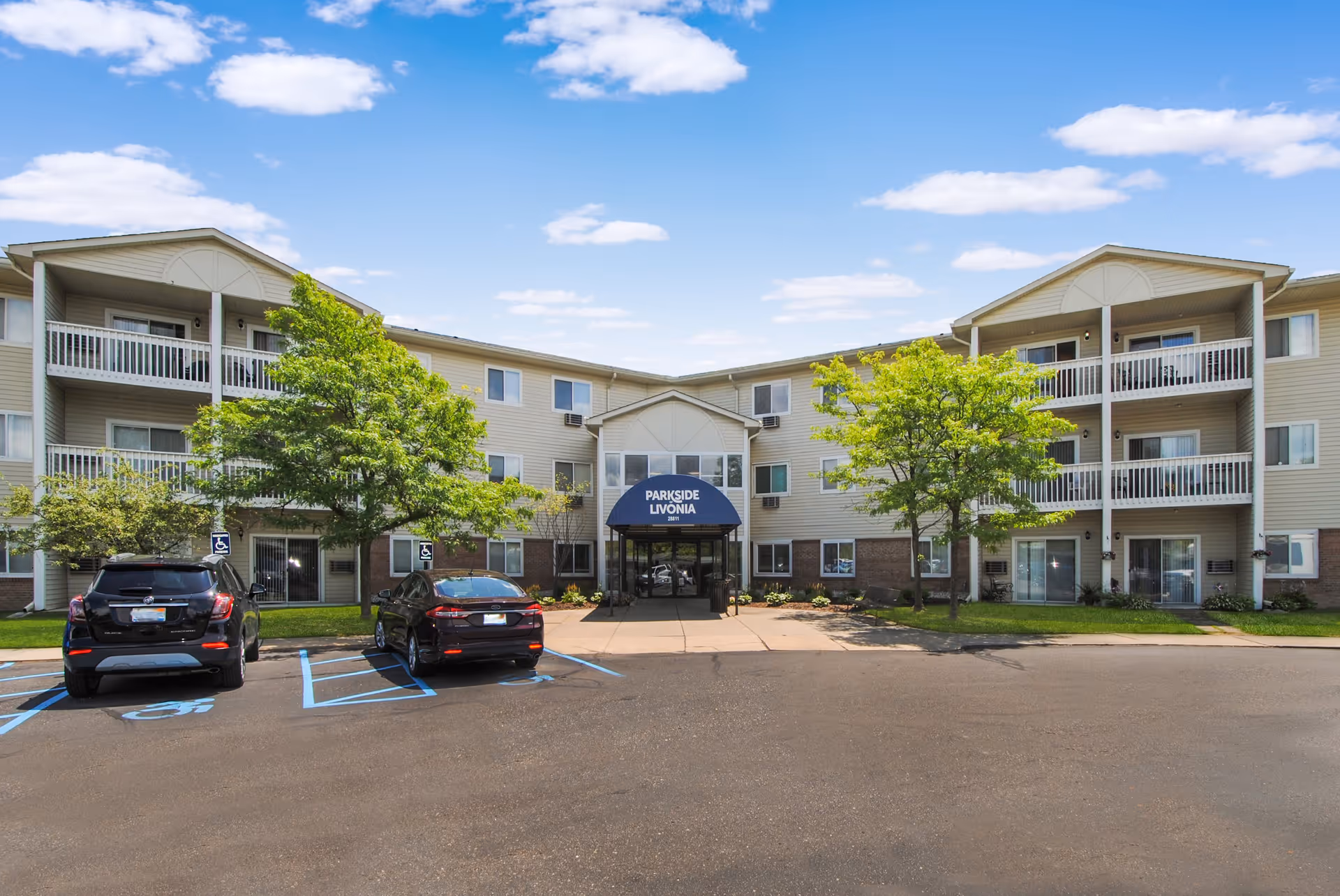 Exterior front view of Parkside of Livonia, a three-story residential building with balconies, trees, and a parking lot with handicap parking spaces under a blue sky with scattered clouds.