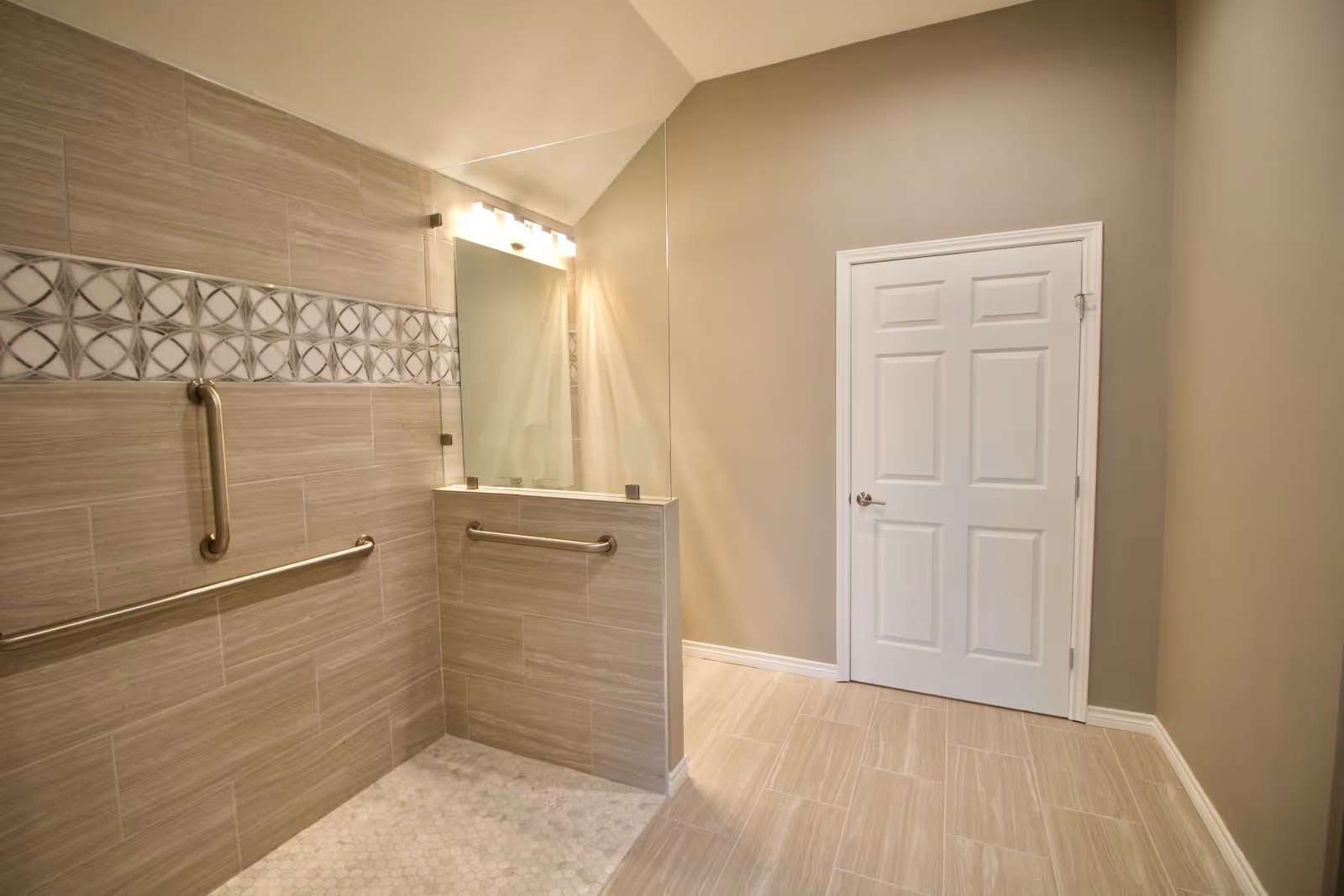 A modern, accessible bathroom featuring a walk-in shower with beige tiles and a decorative tile border. The shower has stainless steel grab bars and a half glass partition. There is a large mirror above a half wall, and a white door is visible on the right side of the room.