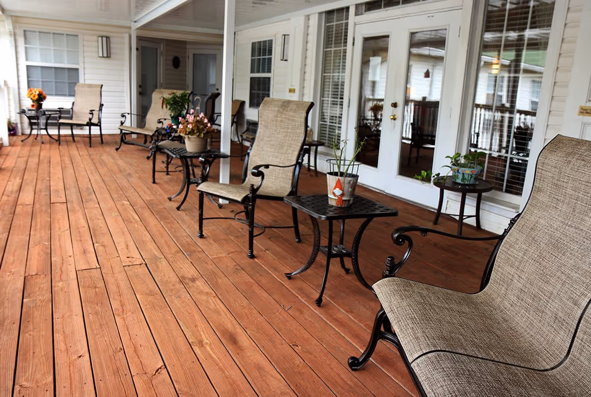 A spacious wooden porch with several cushioned metal chairs and small tables adorned with potted plants and flowers. The porch is attached to a white building with large windows and glass doors, creating a cozy outdoor seating area.