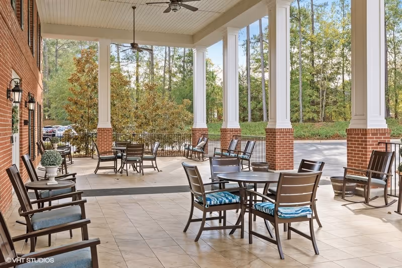 Covered outdoor patio area with several tables and chairs, some with blue cushions, surrounded by white columns with brick bases. Trees and greenery are visible in the background under a clear sky.