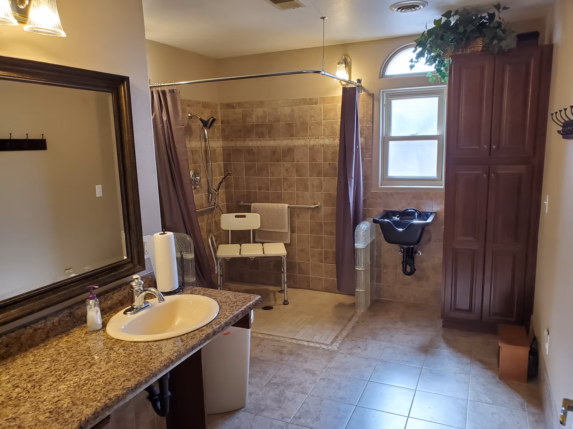 A spacious bathroom with a granite countertop sink, a large mirror, a walk-in tiled shower with a shower chair and grab bars, a small black wall-mounted sink under a frosted window, and a tall wooden cabinet. The room has beige tiled floors and walls, and a shower curtain rod surrounding the shower area.
