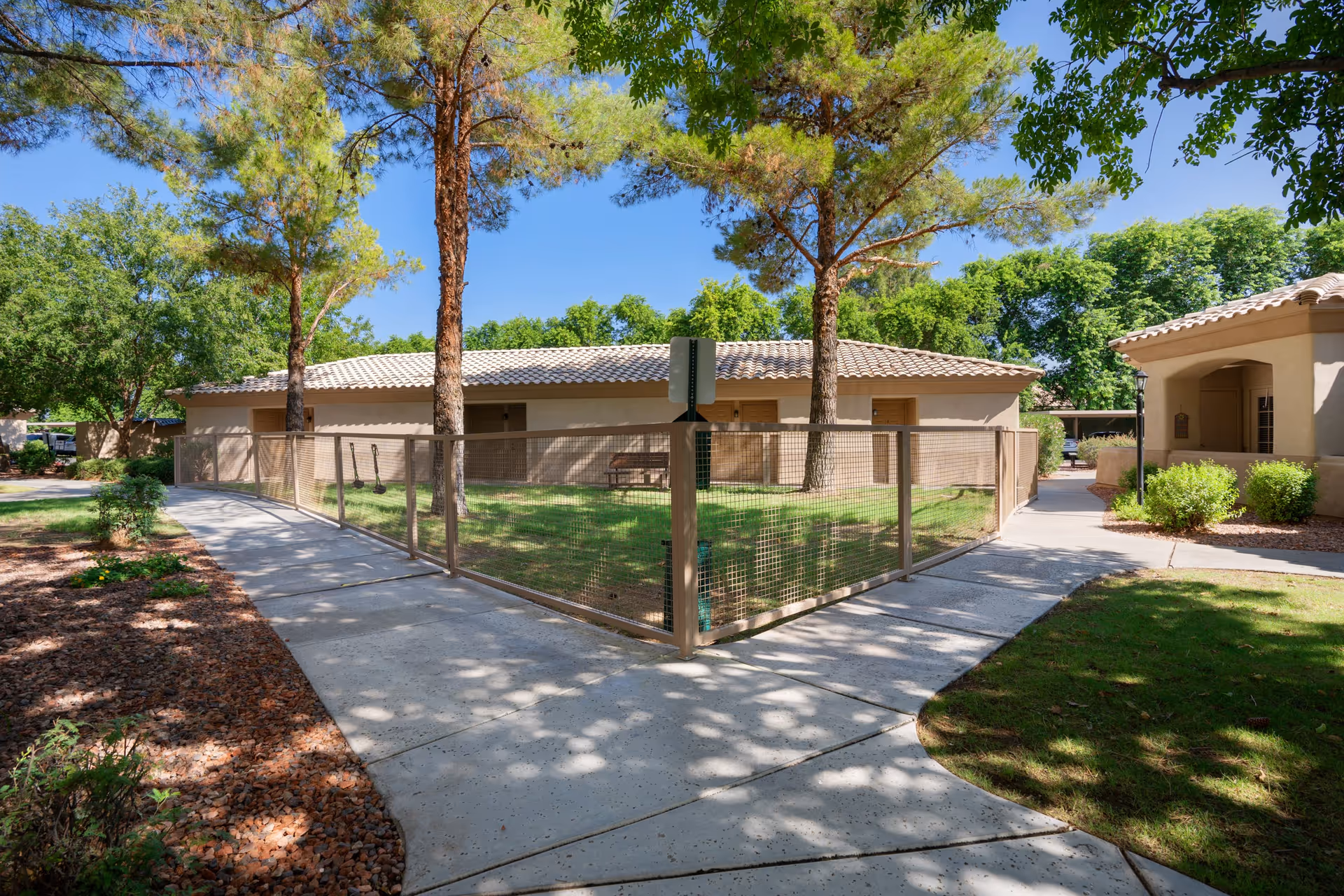 Sunlit courtyard with a fenced grassy area, paved walkways, trees, and single-story stucco buildings.