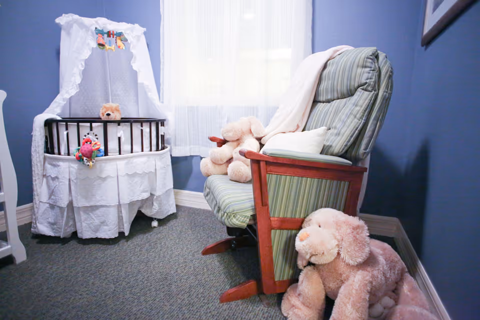 A cozy corner of a room with a white bassinet draped with lace fabric and a teddy bear inside. Next to it is a green striped cushioned rocking chair with a light blanket and stuffed animals placed on and around it. The walls are painted blue and a window with sheer white curtains lets in natural light.