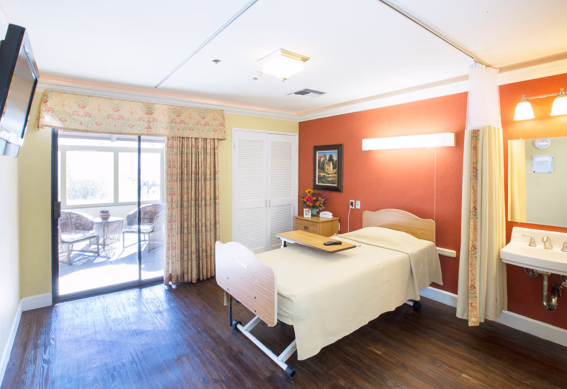A bright and clean senior healthcare bedroom with a single hospital-style bed covered in beige linens. The room features a wooden floor, a bedside table with a flower vase and telephone, a wall-mounted light above the bed, a sink with a mirror, and a sliding glass door leading to a sunlit outdoor seating area with wicker chairs and a table.