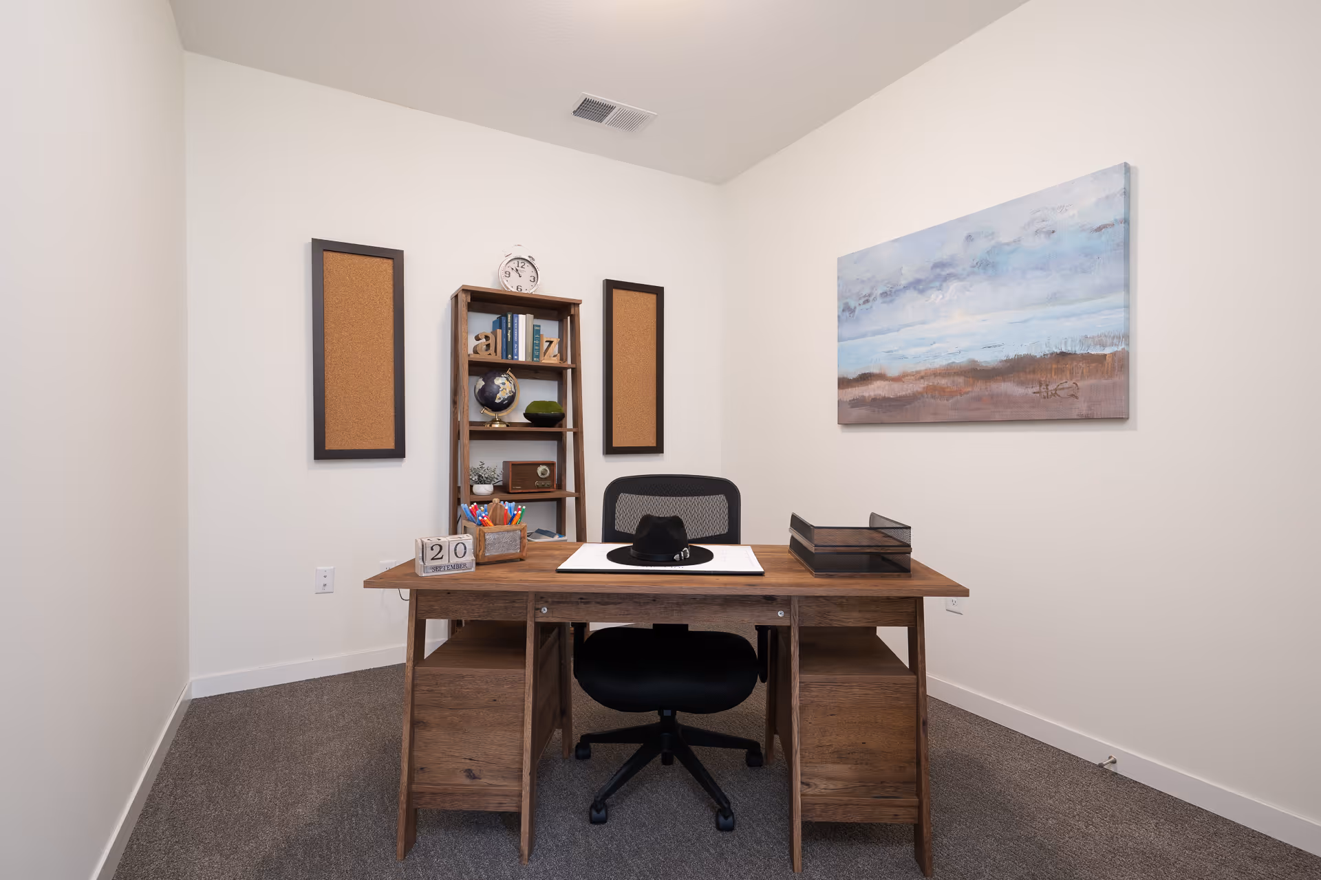 Small office room with a wooden desk, black swivel chair, bookshelf and wall art on white walls.