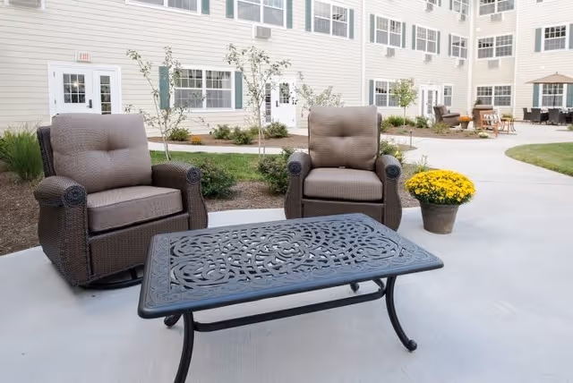Outdoor patio area at The Residence at Glastonbury featuring two cushioned wicker armchairs and a decorative metal table on a concrete surface, with a potted yellow flower and a multi-story building with windows and doors in the background.