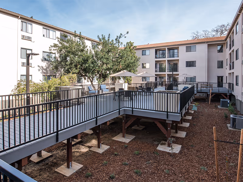 Raised outdoor patio with tables, chairs, and umbrellas in the courtyard of a multi-story senior living building.