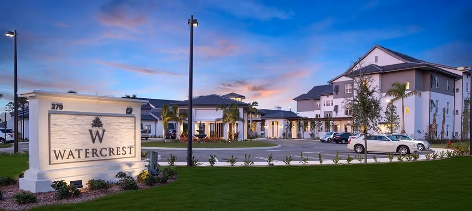 Front exterior of the Watercrest St. Lucie West senior living building and entrance with an illuminated sign and parked cars at dusk.