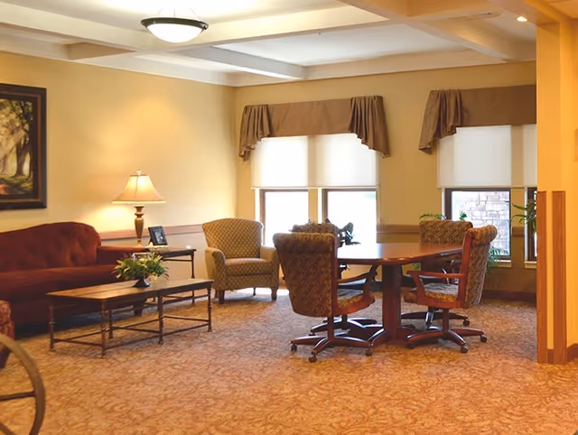 A cozy living room area in a retirement community featuring a red sofa, two armchairs, a wooden coffee table with a flower arrangement, a round wooden table with four cushioned swivel chairs, two windows with brown valances, a table lamp, and a framed painting on the wall.