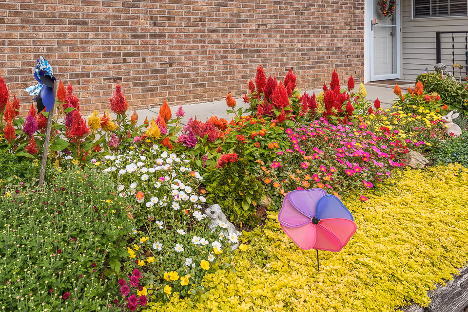 A colorful flower garden with various blooming flowers including red, pink, yellow, orange, and white blossoms in front of a brick wall and a door. The garden features decorative elements such as a purple and pink pinwheel and small animal statues.