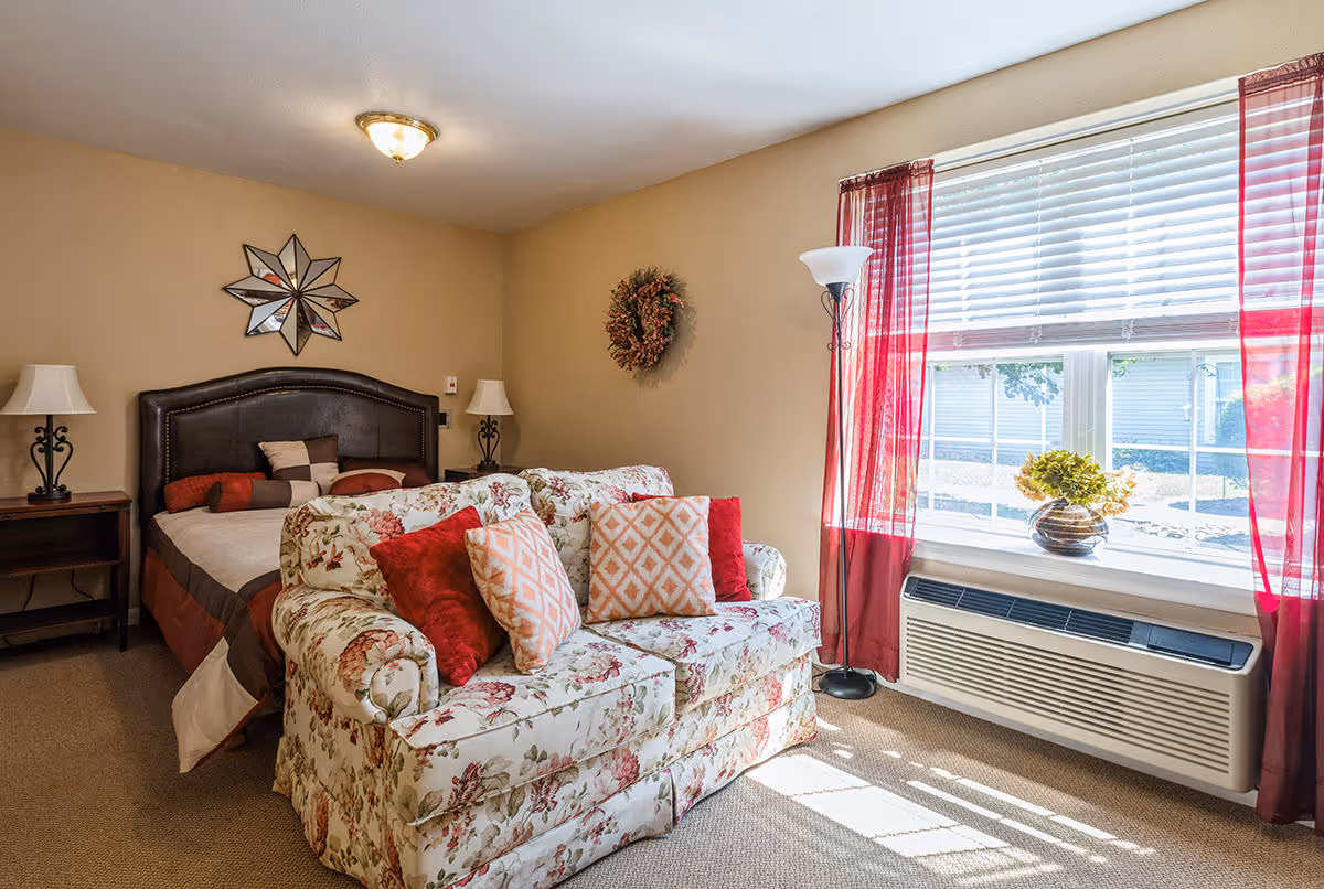 A cozy bedroom with a floral patterned loveseat adorned with red and patterned pillows in front of a bed with a dark headboard and multiple pillows. The room has beige walls, a window with red sheer curtains letting in sunlight, a floor lamp, two bedside tables with lamps, a decorative wreath on the wall, and a star-shaped mirror above the bed.