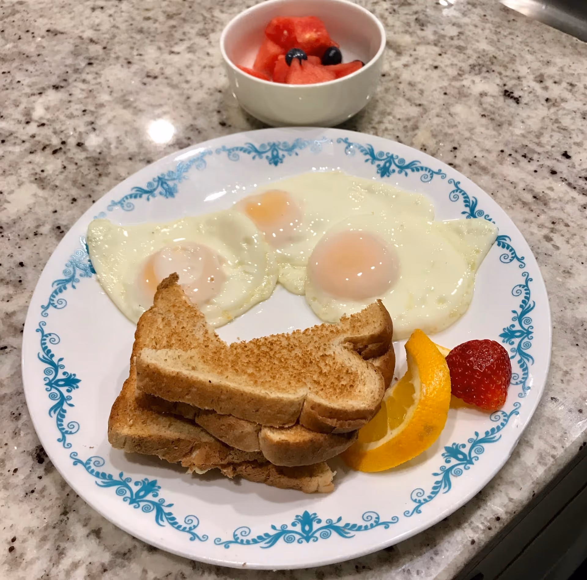 A plate with three sunny-side-up eggs, three slices of toasted bread, a slice of orange, and a strawberry on a granite countertop. Behind the plate is a small white bowl containing pieces of watermelon and blueberries.