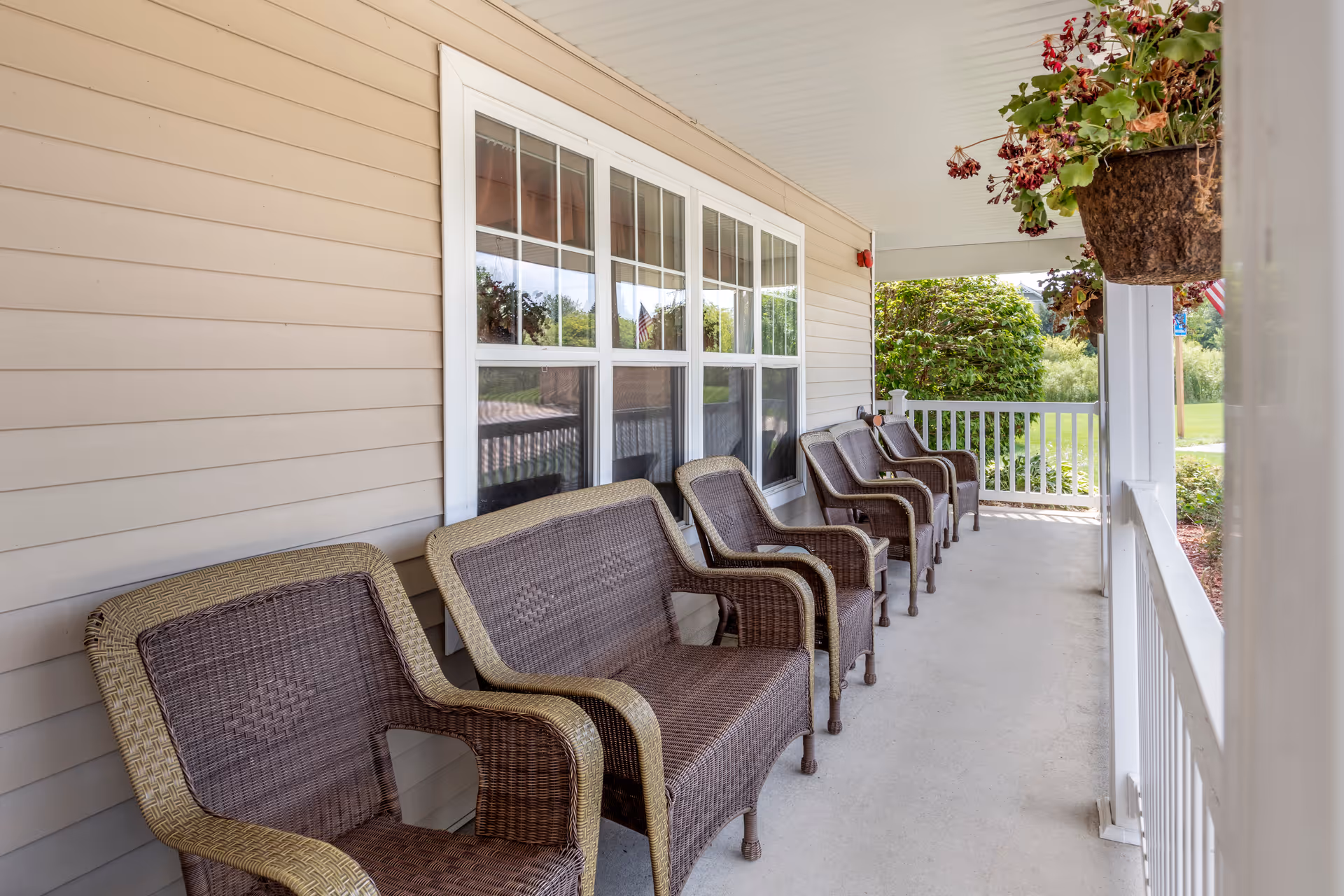 A covered outdoor porch area with a row of six brown wicker chairs lined up against the beige siding of a building. There are white-framed windows behind the chairs and a hanging basket with plants on the right side. Green bushes and a lawn are visible beyond the white railing of the porch.