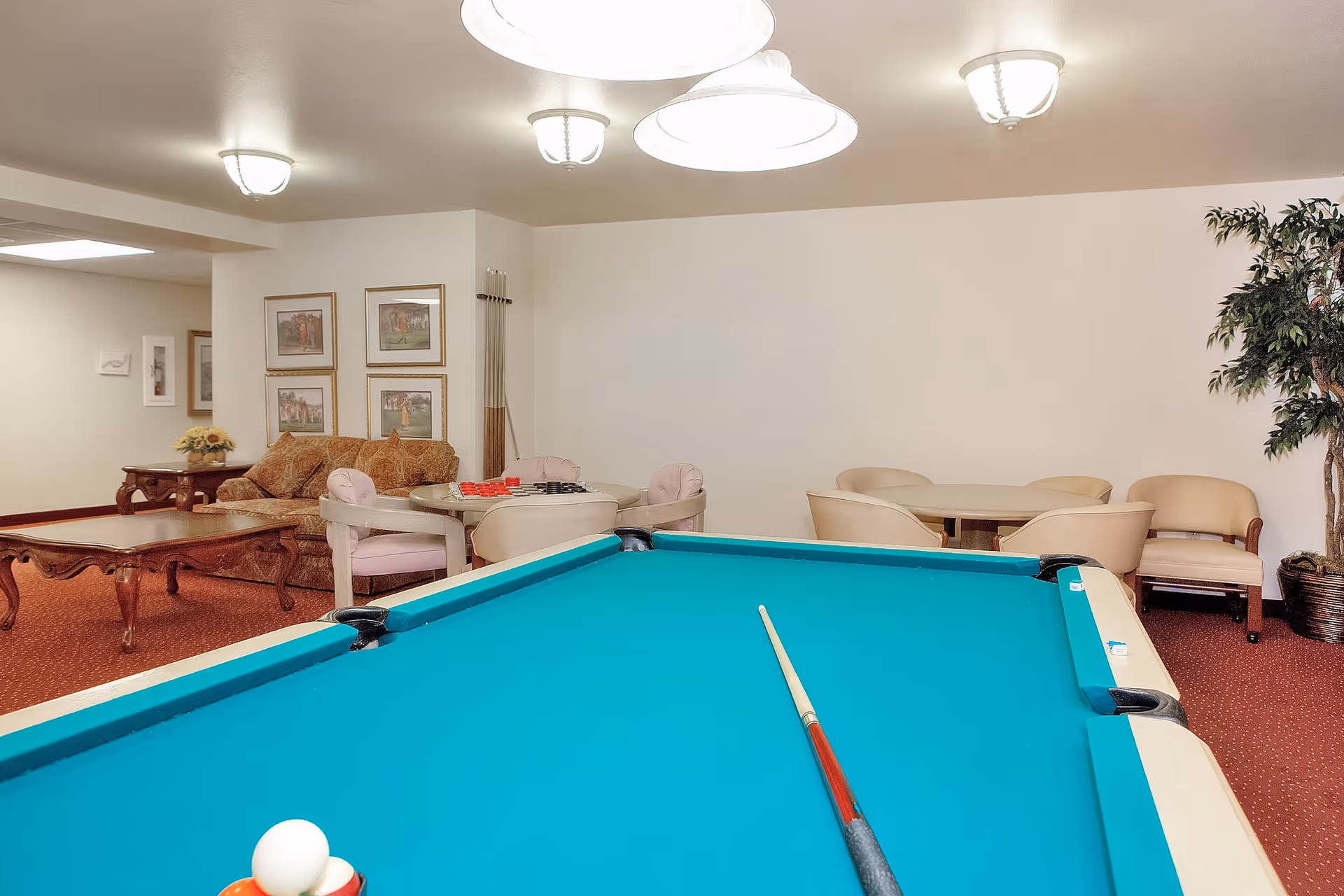 Interior view of a recreational room with a pool table in the foreground, a round table with chairs and a checkers game, a sofa with framed pictures on the wall behind it, and additional seating with beige chairs and a potted plant in the corner.