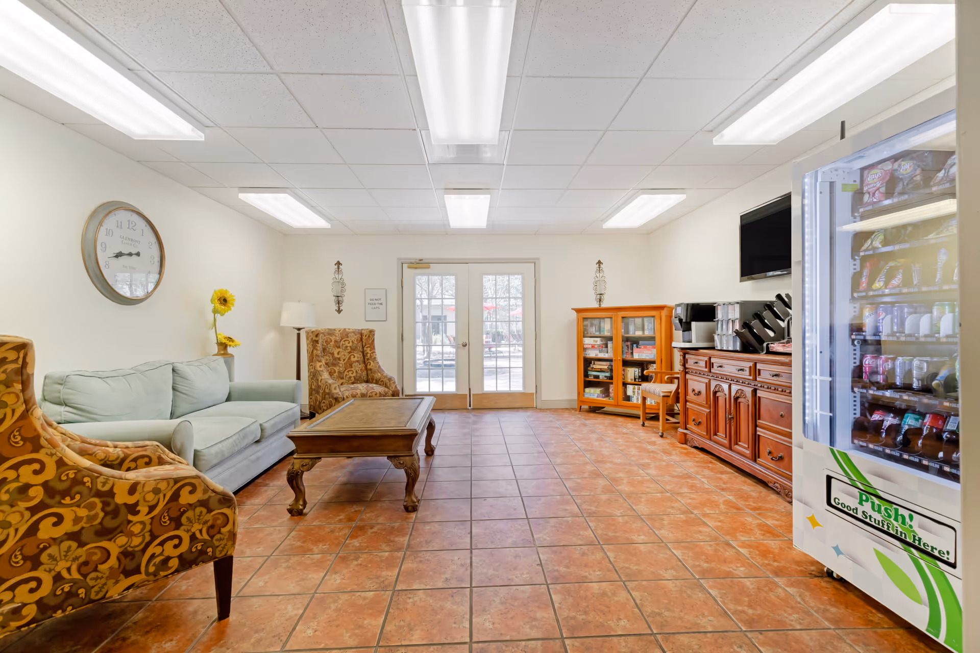 A well-lit common room with terracotta tiled floor, featuring a light green sofa and two patterned armchairs around a wooden coffee table. On the right side, there is a vending machine stocked with snacks and drinks, a wooden cabinet with a coffee machine, and a glass-fronted cabinet with board games. Double glass doors lead outside, and a wall clock and a floor lamp are visible on the left wall.