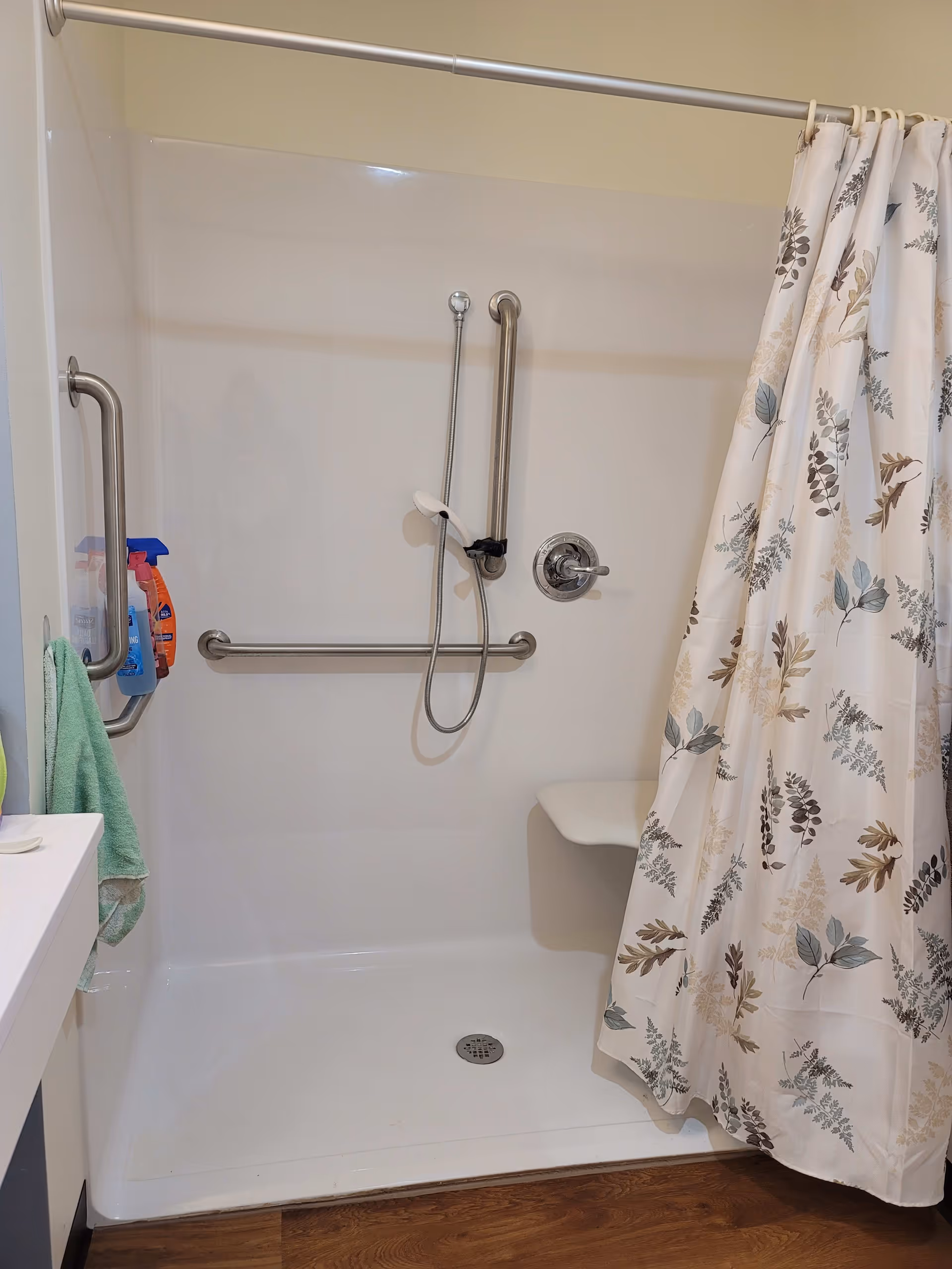 A walk-in shower with a white base and walls, equipped with stainless steel grab bars on the side and back walls. There is a handheld showerhead attached to the wall, and a floral-patterned shower curtain partially drawn to the right. A small built-in seat is visible in the shower corner. Several cleaning products and a green towel hang on the left grab bar. The floor outside the shower is wood.