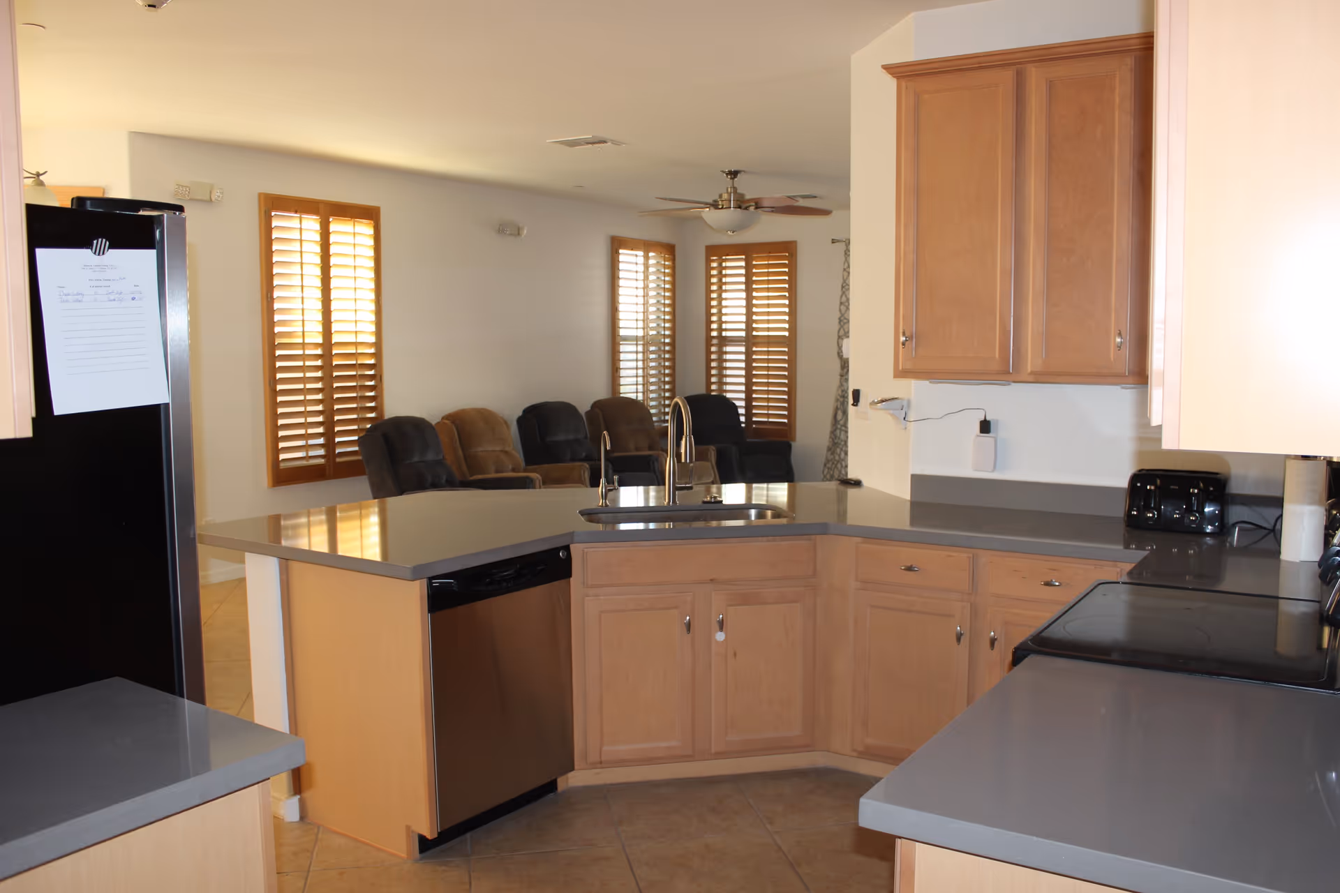 A modern kitchen with light wood cabinets and gray countertops. The kitchen features a stainless steel dishwasher, a black stove, a black refrigerator with a paper attached, and a double sink with a high-arc faucet. In the background, there is a living area with several recliner chairs and windows with wooden shutters.