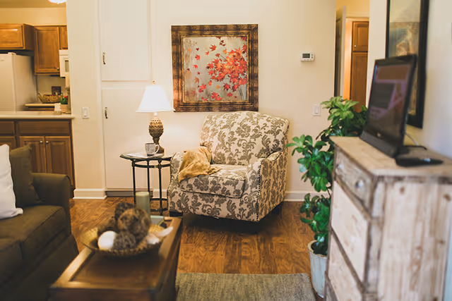 Cozy living room area with a patterned armchair, a side table with a lamp and a cup, a wooden coffee table with decorative items, a sofa with a white pillow, a wooden dresser with a TV on top, and a view into a kitchen with wooden cabinets.