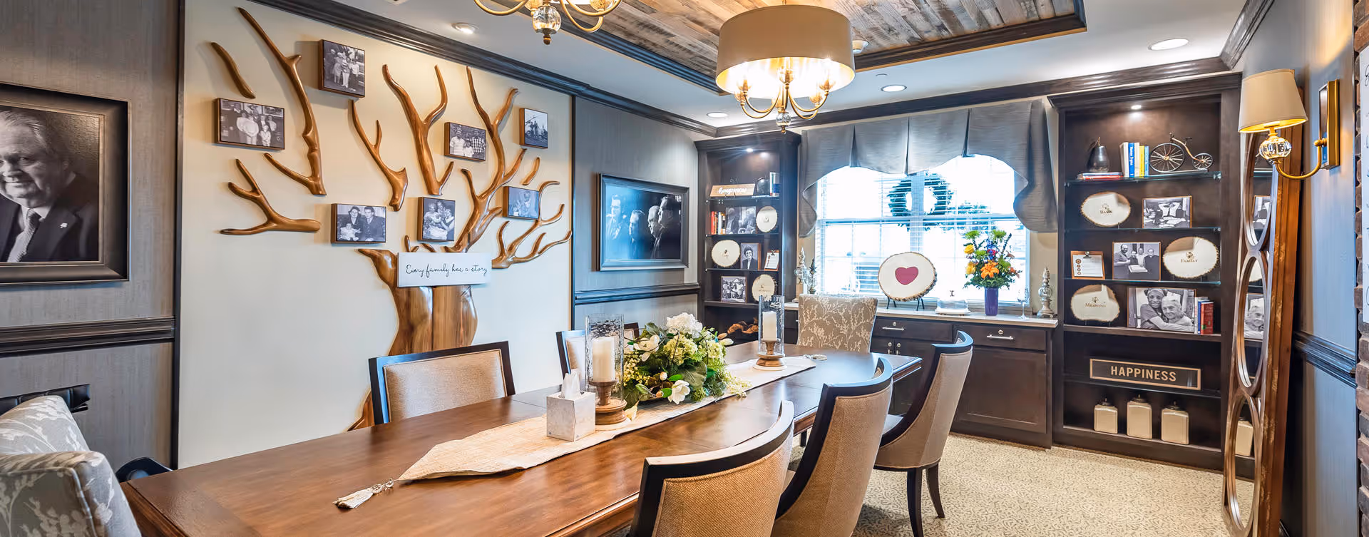 A warmly decorated dining room with a long wooden table surrounded by cushioned chairs. The walls feature framed black and white photographs and a wooden tree-shaped wall art with photos attached. There are built-in shelves with decorative plates, books, and flowers near a window with blinds. The room has a cozy and elegant ambiance with soft lighting from ceiling and wall fixtures.