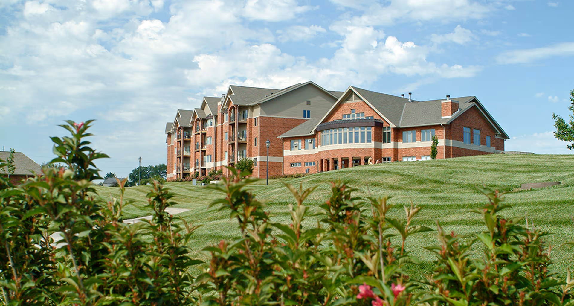 A large brick senior living facility building with multiple floors and balconies, situated on a grassy hill with a partly cloudy sky in the background. Green shrubs with some pink flowers are visible in the foreground.