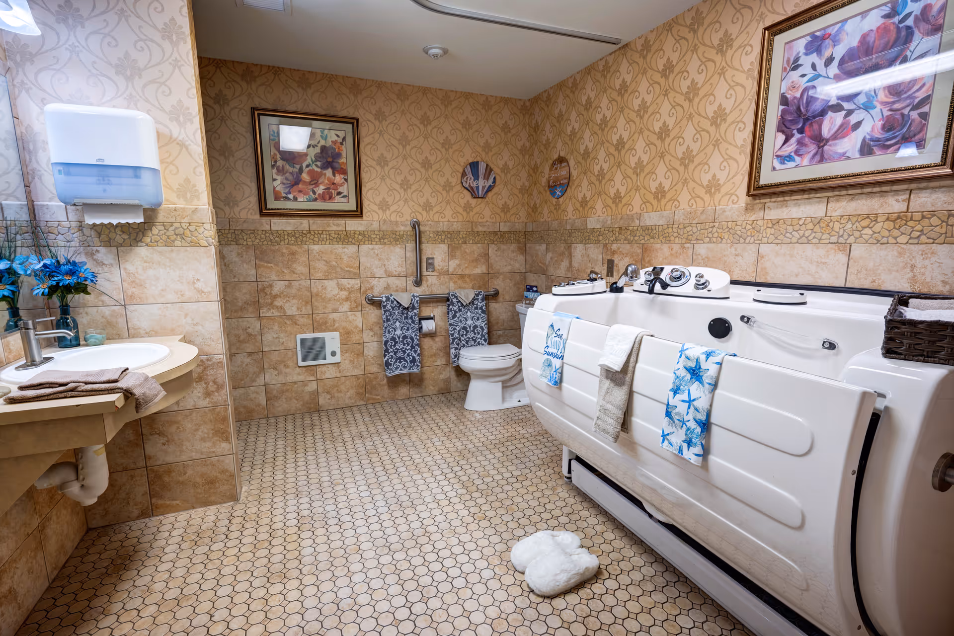A spacious bathroom with beige patterned tile walls and floor. The room features a white sink with a towel and blue flowers on the left, a toilet with two patterned towels hanging on a rail above it, and a large white walk-in bathtub with several towels draped over its side. The walls are decorated with framed floral artwork and small decorative plaques. A pair of white slippers is placed on the floor in front of the bathtub.
