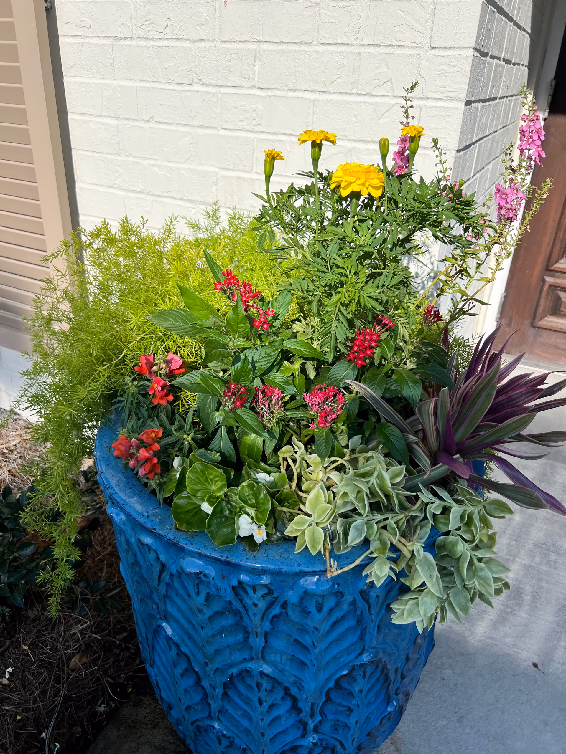 A large blue decorative planter filled with a variety of colorful flowers and green plants, placed outside near a white brick wall and a wooden door.
