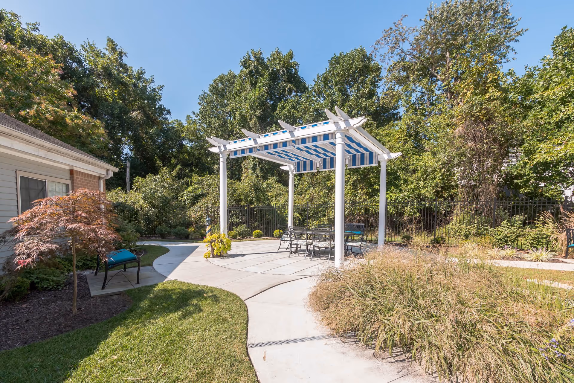 Outdoor patio area at Lighthouse Senior Living at Ellicott City featuring a white pergola with a blue and white striped canopy, metal table and chairs underneath, surrounded by greenery, trees, and landscaped garden beds with a curved concrete walkway.