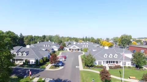Aerial view of Tennyson Court senior living facility showing multiple single-story buildings with gray roofs arranged around a central driveway. The area is surrounded by green lawns, trees, and parked cars under a clear blue sky.