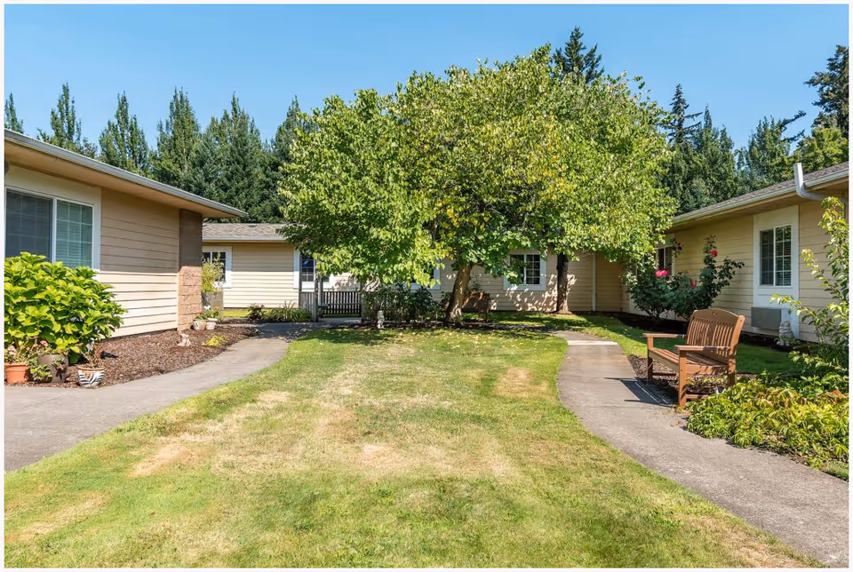 A sunny outdoor courtyard area at The Cottages Senior Living Memory Care featuring a grassy lawn, paved walkways, a wooden bench, various plants and shrubs, and beige single-story buildings surrounding the space with trees in the background.