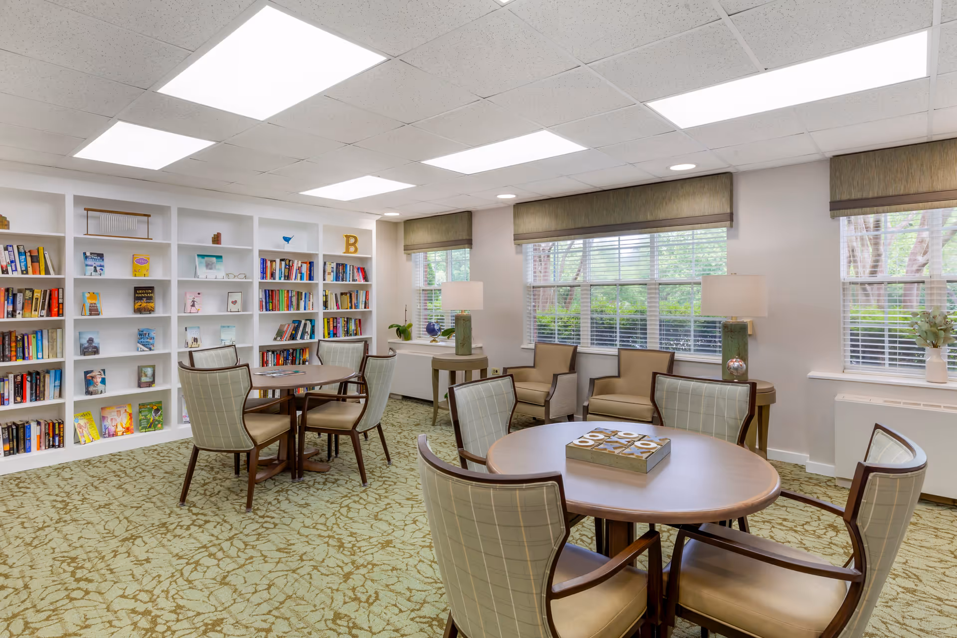 A bright and cozy common area with round wooden tables and cushioned chairs arranged for socializing or reading. The room features large windows with blinds, two table lamps on side tables, and built-in white bookshelves filled with books and decorative items. The carpet has a green pattern, and the ceiling has recessed lighting panels.