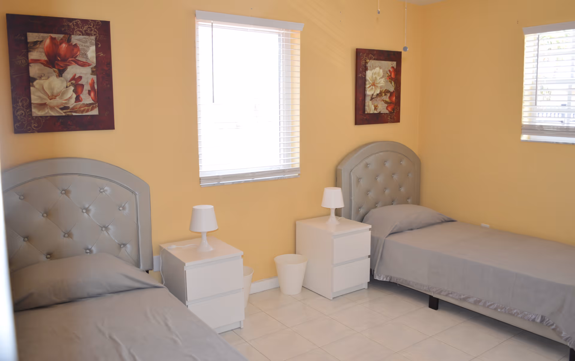 A bedroom with two single beds featuring gray tufted headboards and gray bedding. Each bed has a white nightstand beside it with a small white lamp. The walls are painted yellow and decorated with floral paintings above each bed. Two windows with white blinds allow natural light into the room. The floor is tiled in white.