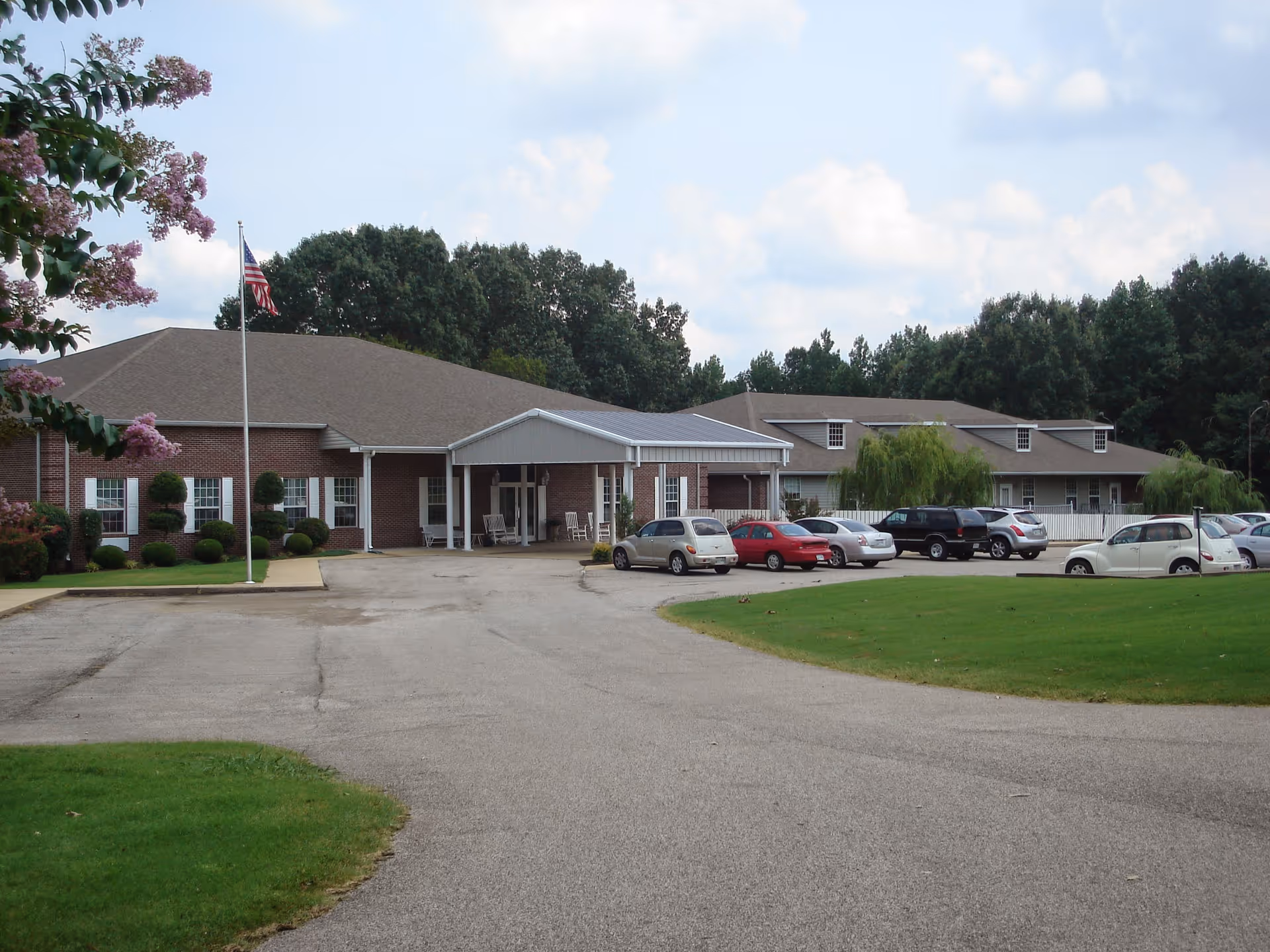 Exterior view of a single-story brick building with a covered entrance and several parked cars in front. The building is surrounded by green lawns, trees, and a flagpole with an American flag. The sky is partly cloudy.