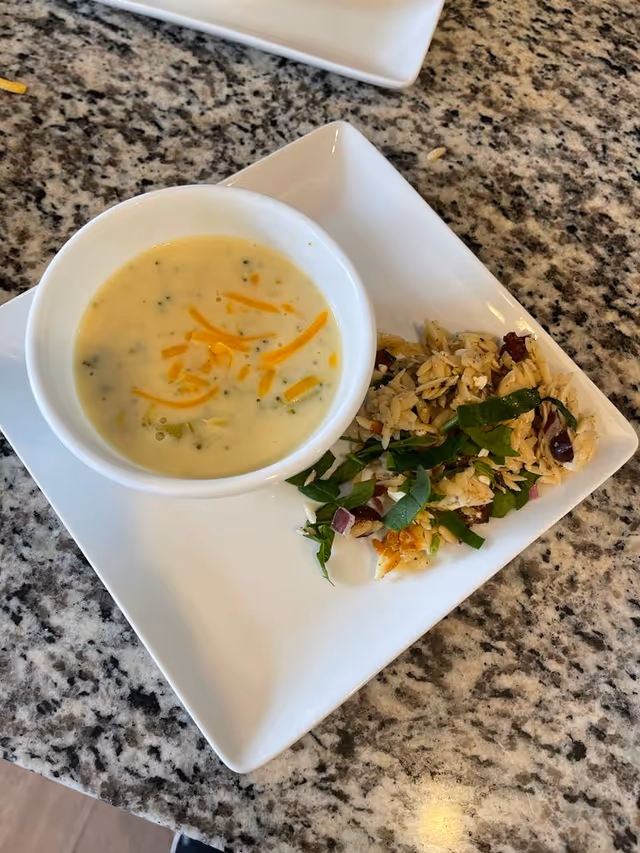 A white bowl of creamy soup topped with shredded cheddar cheese, served on a white square plate alongside a small portion of orzo pasta salad with greens and dried cranberries, placed on a granite countertop.