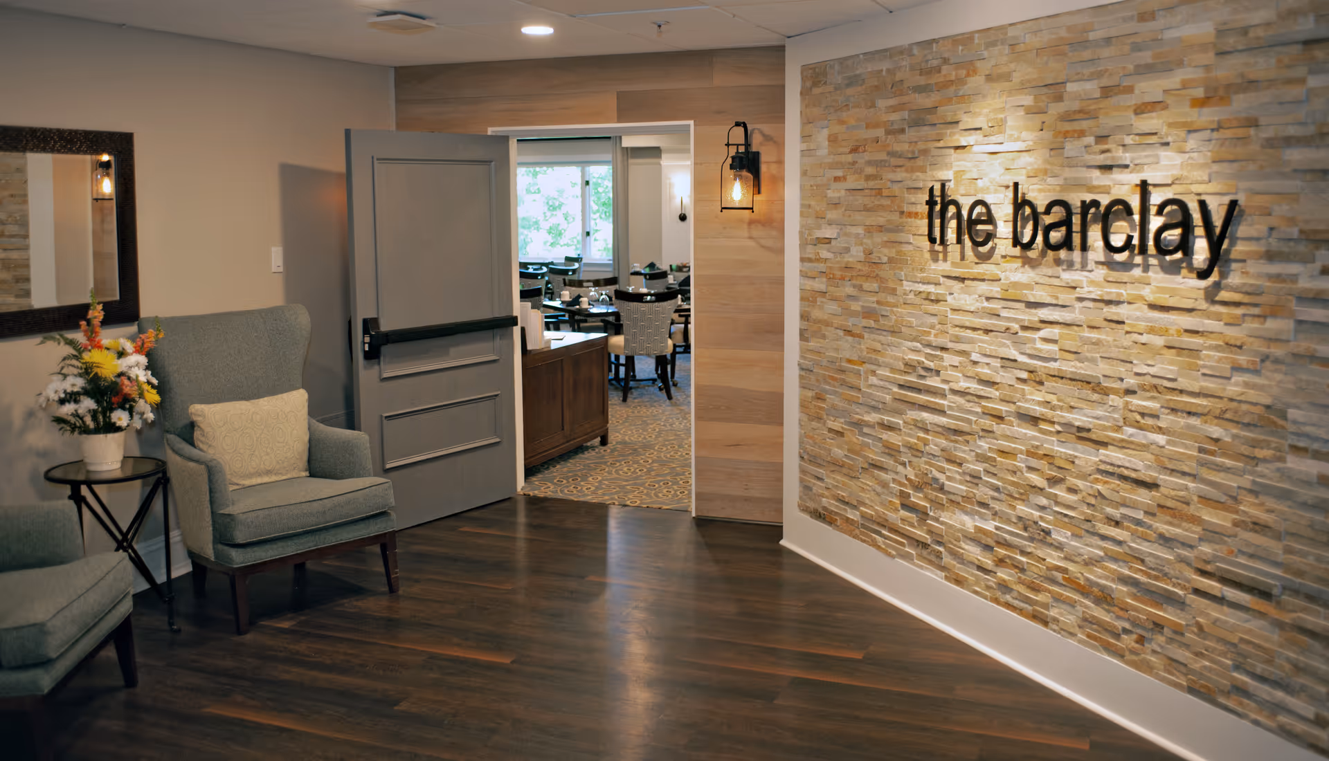Interior view of a senior living facility hallway with wooden flooring, two upholstered armchairs with cushions, a small round table with a flower arrangement, a large mirror on the wall, and an open door leading to a dining area. The wall on the right features a textured stone design with the words 'the barclay' mounted on it.