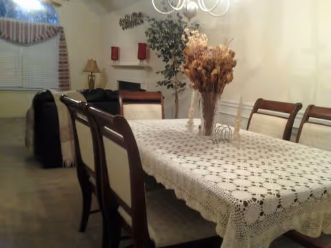 A dining room with a rectangular table covered by a white lace tablecloth. Six wooden chairs with cushioned seats surround the table. A vase with dried flowers is placed in the center of the table. In the background, there is a living area with a black sofa, a lamp, a fireplace with red candles on the mantel, a large potted plant, and a window with a striped valance.