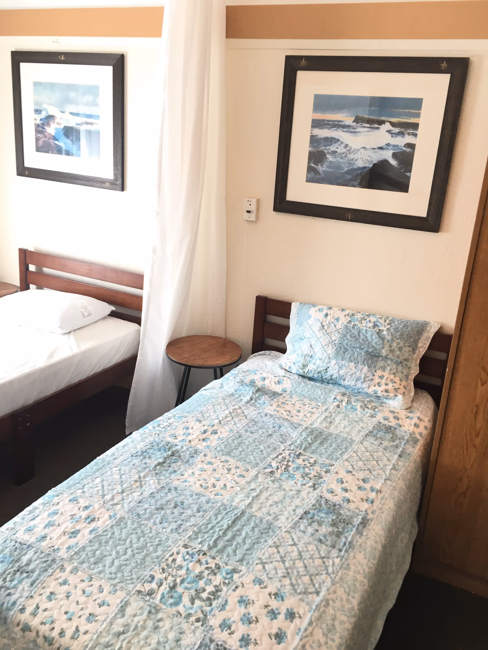 A small bedroom with two single beds. One bed is made with a blue and white patchwork quilt and matching pillow, while the other bed has a plain white sheet and pillow. Between the beds is a small round wooden side table. Above each bed hangs a framed painting depicting ocean scenes. A white curtain is partially visible between the beds.