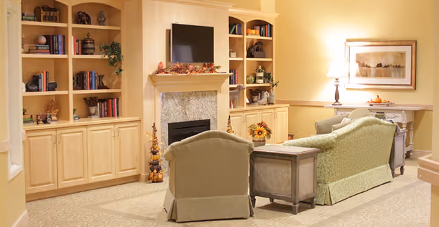 A cozy living room with light yellow walls, a stone fireplace with a mantel decorated with autumn leaves, built-in wooden shelves filled with books and decorative items, a flat-screen TV mounted above the fireplace, a green upholstered sofa and armchair, a wooden side table, a lamp on a console table, and a framed landscape painting on the wall.