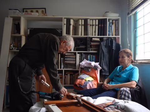 Two people in a small furnished room; one person is working on a wooden project on a bed while the other sits nearby next to a bookshelf and window.