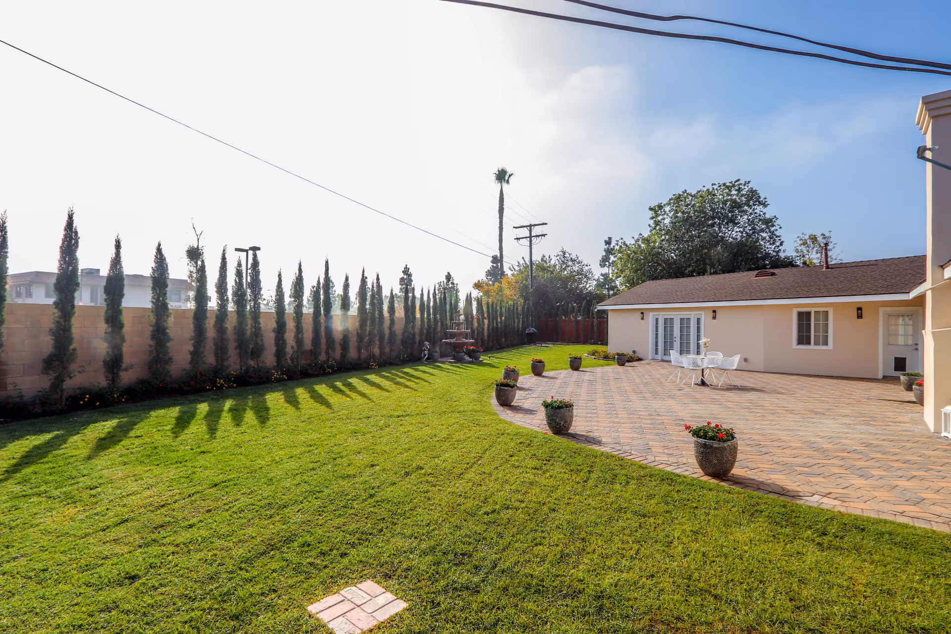 A spacious outdoor patio area with a tiled floor and several potted plants. There is a white table with chairs on the patio. The patio is adjacent to a beige building with white doors and windows. A well-maintained green lawn extends from the patio, bordered by a row of tall, narrow trees along a brick wall. The sky is clear with some light clouds.