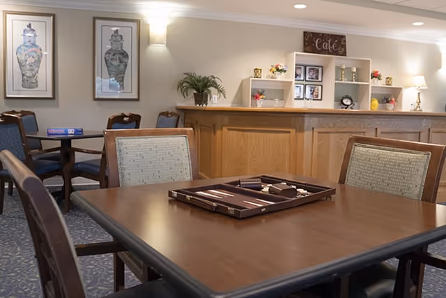 Interior view of a senior living facility common area with wooden tables and cushioned chairs. A backgammon game is set up on the nearest table. The background features a wooden counter with shelves holding decorative items, framed pictures, and a sign that reads 'Cafe'. The walls are adorned with framed artwork and soft lighting fixtures.