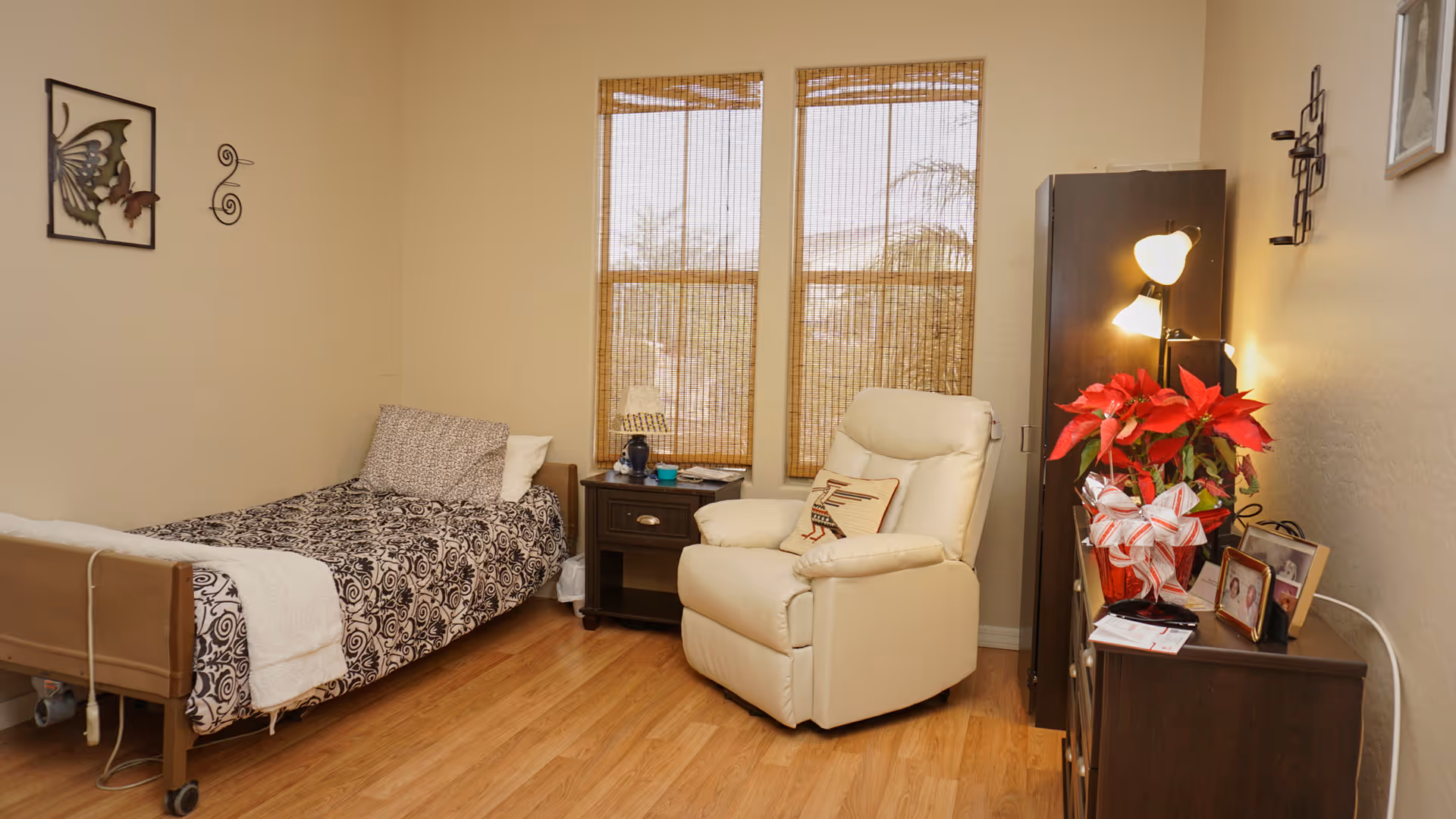 A cozy senior living bedroom featuring a single bed with patterned bedding and a white towel, a wooden nightstand with a lamp and small items, a comfortable cream-colored recliner chair with a decorative pillow, two windows with bamboo blinds, and a wooden dresser adorned with a red poinsettia plant, framed photos, and a lamp.
