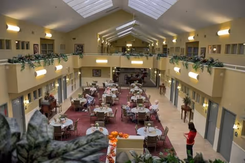 Interior view of a spacious dining area in an assisted living facility with multiple round tables and chairs arranged on a red carpet. The room has high ceilings with skylights, beige walls, and upper-level balconies with plants. A person in a red shirt is standing near the right side of the room.